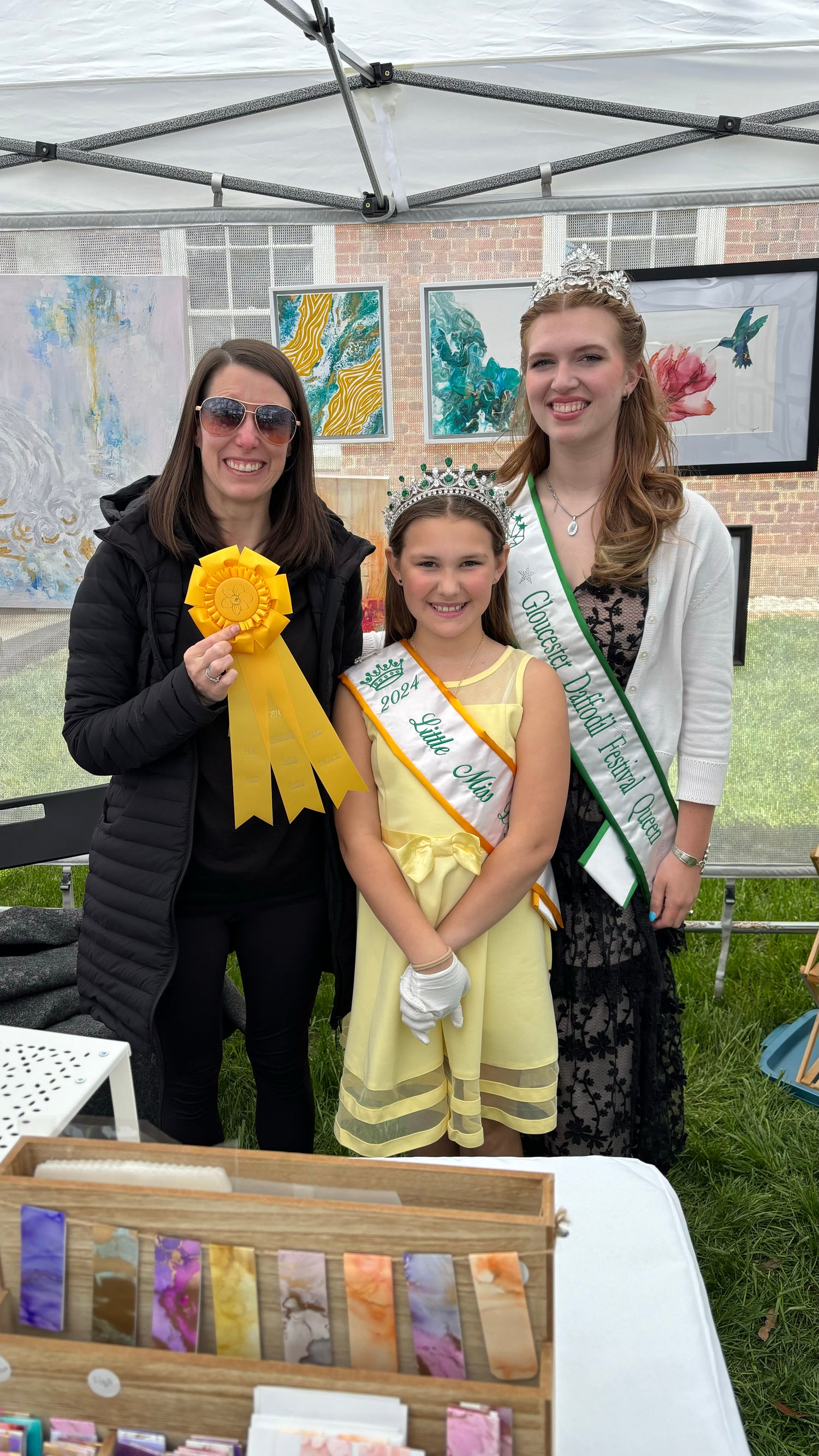 Three women, with the award winner of fine art at the daffodils festival, Josie.Ane.Sakura standing under a tent with art displays in the background.